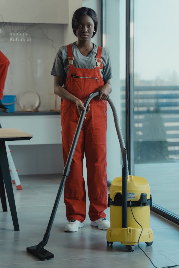 An adult woman in red coveralls operates a vacuum cleaner in a modern indoor setting, focusing on cleaning tasks.